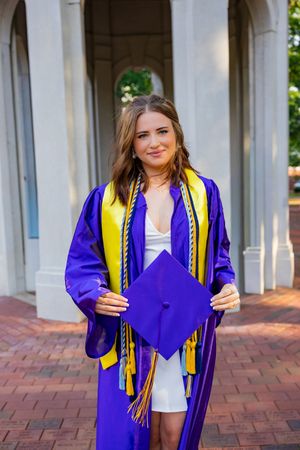 Young woman in purple and gold graduation cap and gown holding mortarboard on brick campus walkway.