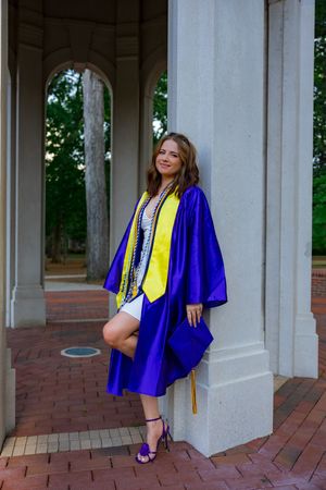 Young woman in purple graduation gown and yellow stole posing confidently between white columns on brick walkway.