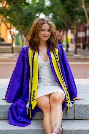 Young woman in purple graduation gown with yellow stole sits on steps, smiling at camera outdoors.