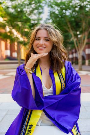 Smiling young woman in purple and yellow graduation gown poses outdoors on campus brick walkway.