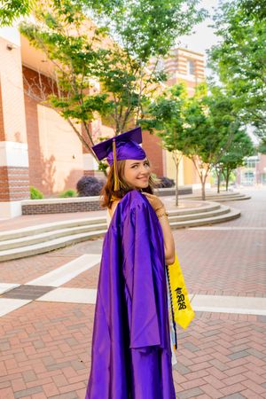 Smiling graduate in purple cap and gown holding a 2026 stole, posing outdoors on a brick walkway near a university building.