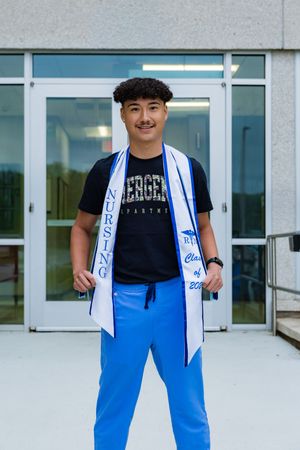 Young male nursing graduate smiles proudly wearing blue scrubs and a white nursing graduation sash outdoors.