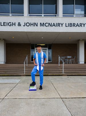 Graduate in blue outfit with stole stands proudly before Leigh & John McNairy Library holding textbooks.
