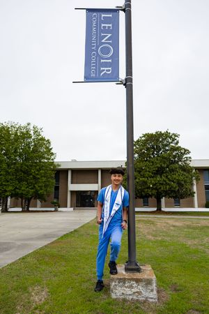 Student in blue scrubs poses by Lenoir Community College banner sign on campus grounds.