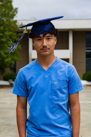 Young man in blue scrubs and graduation cap poses outdoors, celebrating academic achievement in healthcare.