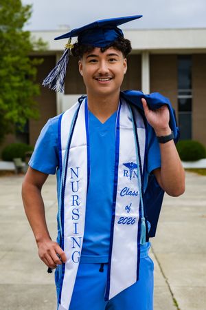 Smiling male nursing graduate in blue scrubs and cap with Class of 2026 graduation stole posing outdoors.