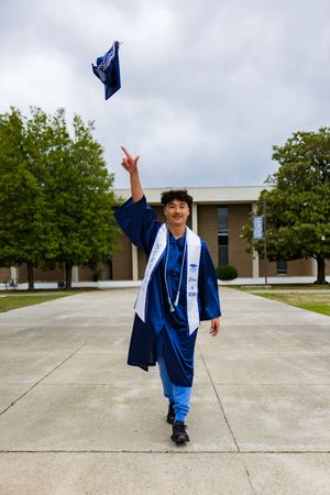 A smiling graduate in blue cap and gown tosses their cap into the air outdoors on a campus walkway.