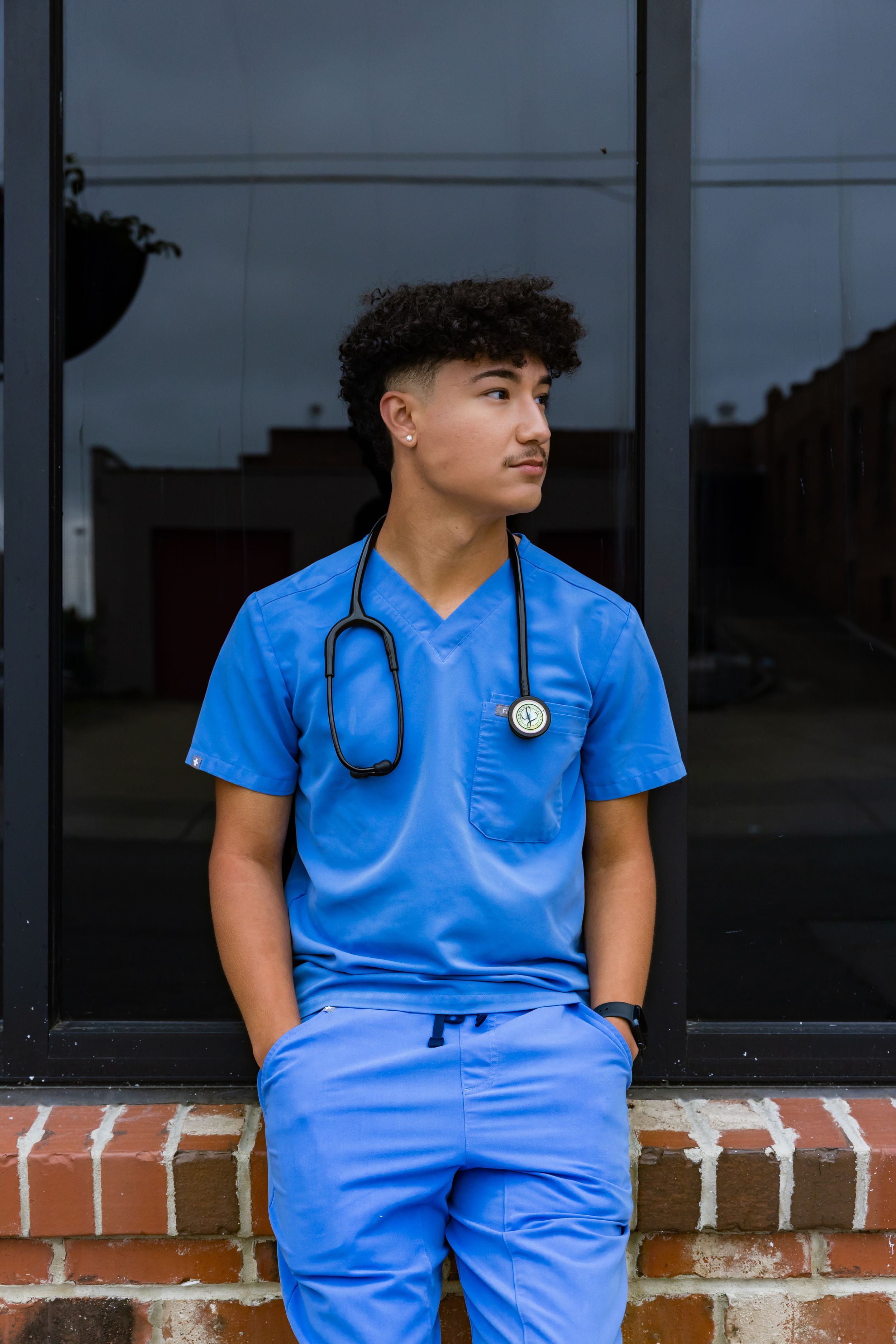 Young male nurse in blue scrubs with stethoscope sitting outdoors against a dark window wall.