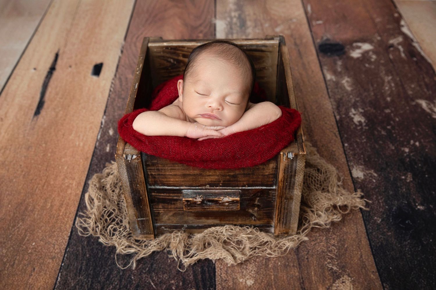 Sleeping newborn baby posed in a rustic wooden crate with red fabric lining for a professional newborn photo.