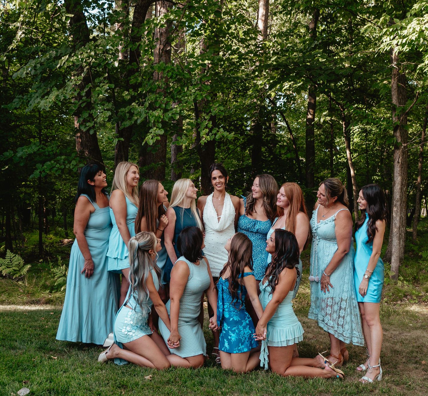 Bride in white dress surrounded by bridesmaids in light blue dresses laughing outdoors in a lush green wooded setting.
