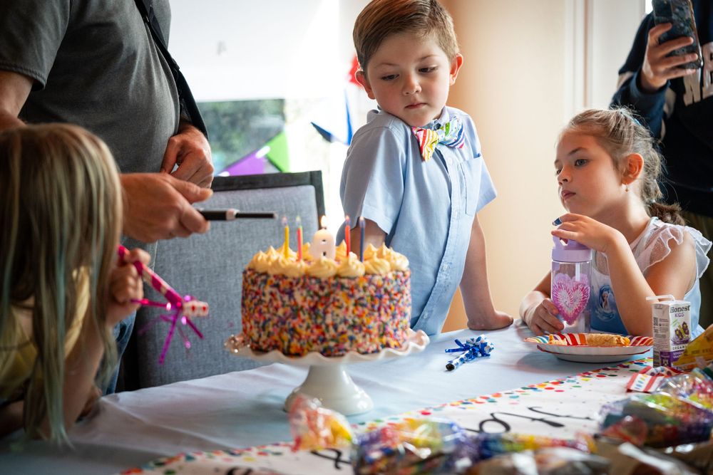 Children gather around a decorated birthday cake at a party with confetti scattered on the table.