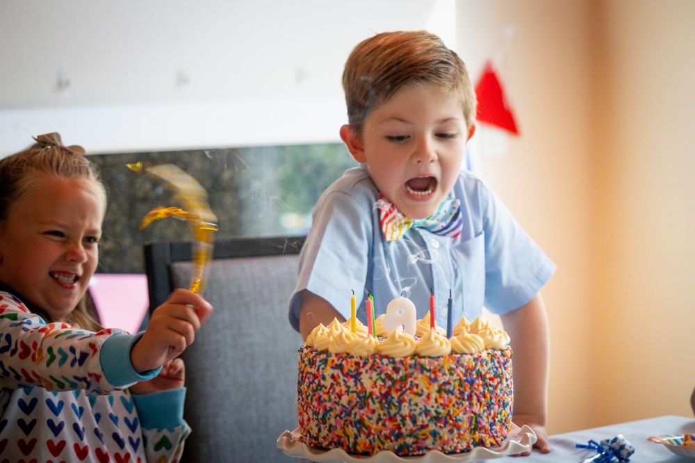 A child blows out birthday candles on a colorful sprinkle-covered cake while another child watches and laughs.