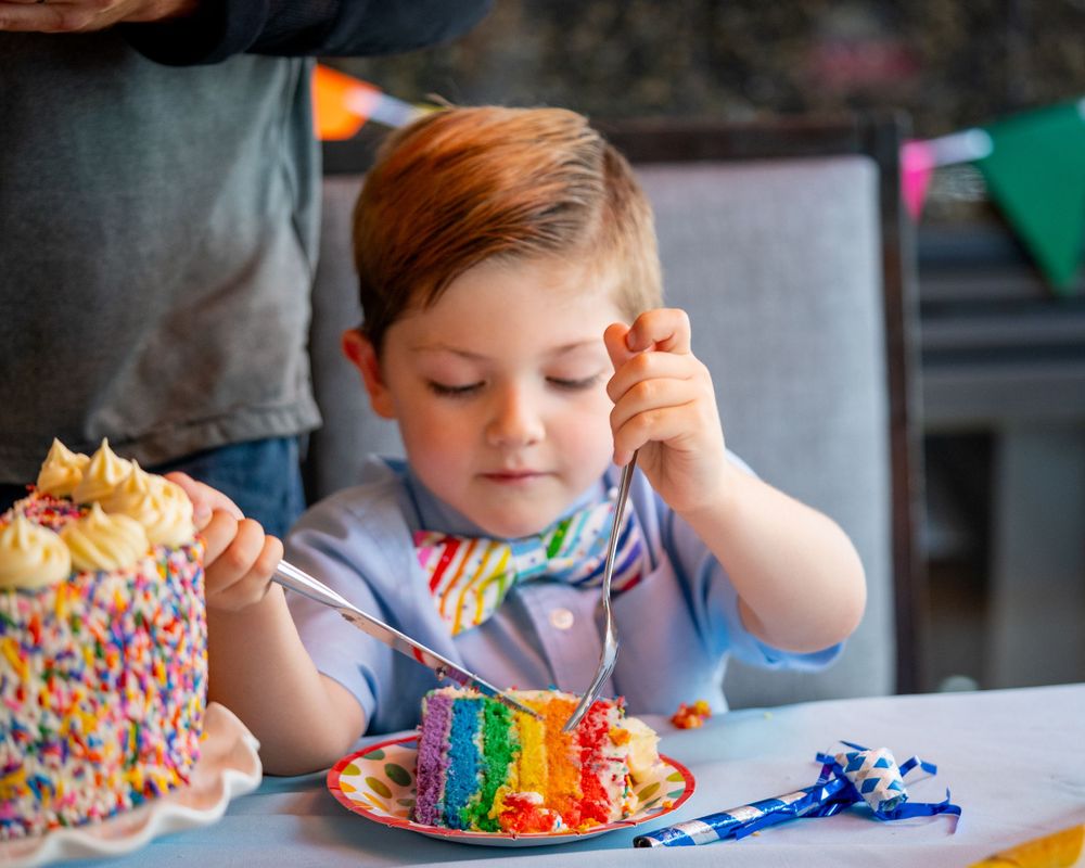 Child enjoying colorful rainbow cake slice covered in sprinkles at a birthday celebration.