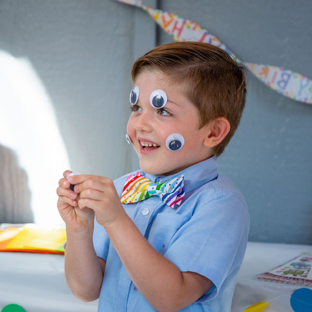 Young child wearing a light blue shirt and googly eye decorations smiles during a creative activity.
