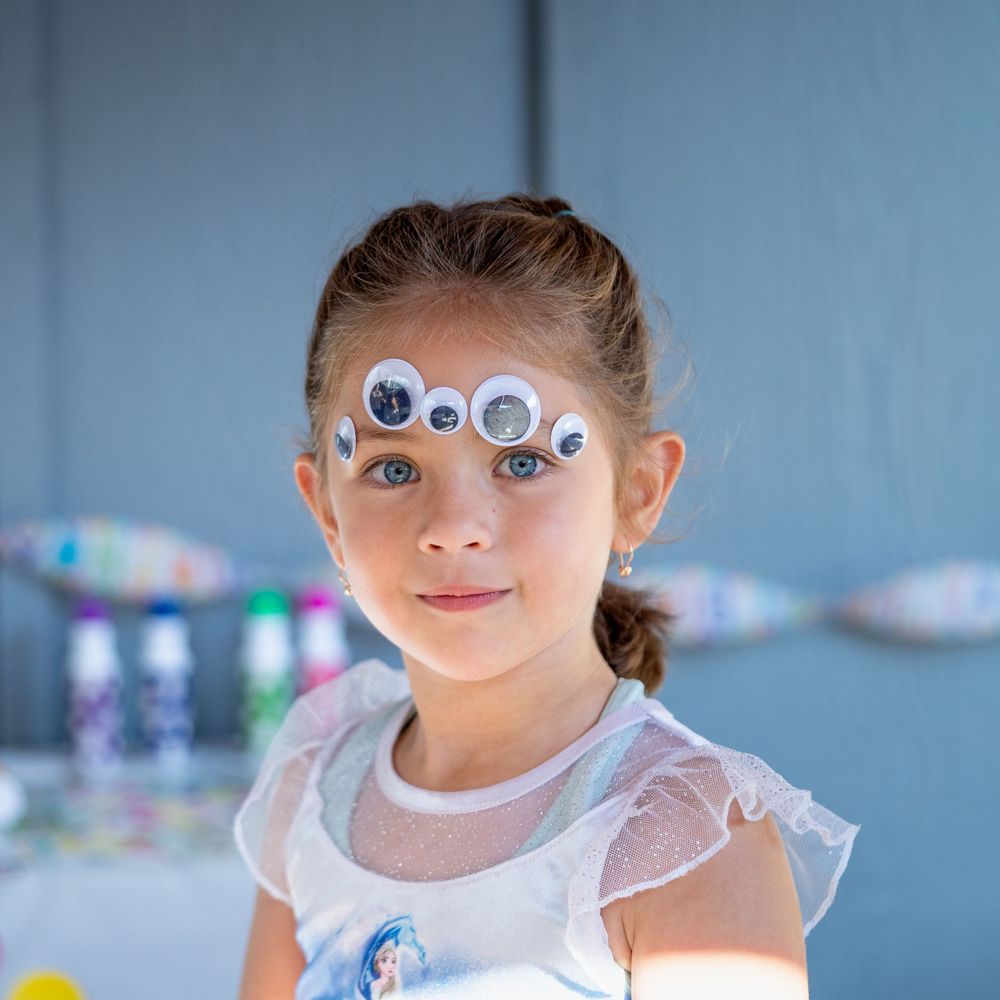 Young child wearing decorative googly eyes on forehead and white ruffled dress stands against blue background.