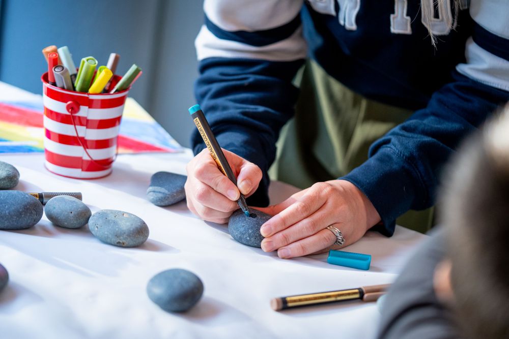 Someone writes on paper with colorful markers in a striped cup holder and smooth gray stones arranged on a white surface.