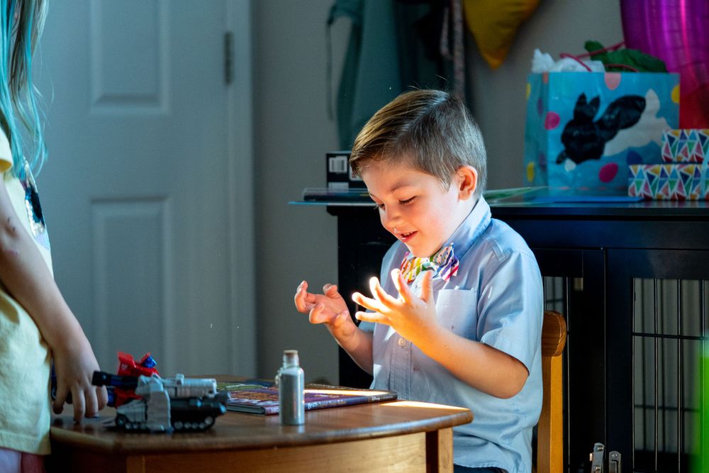 A child in a light blue shirt sits at a table working intently on a small object in natural lighting.