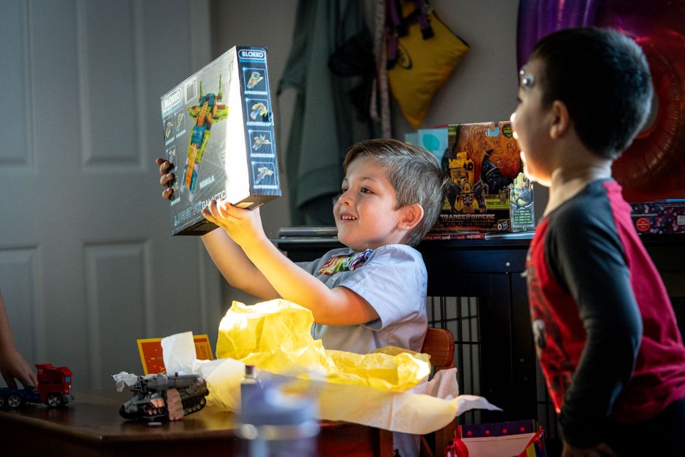 Two children sitting at a table reading a book together in a room illuminated by dramatic window light.