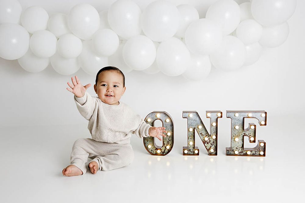 Smiling baby boy in beige knit outfit sitting beside illuminated ONE marquee letters with white balloons backdrop.