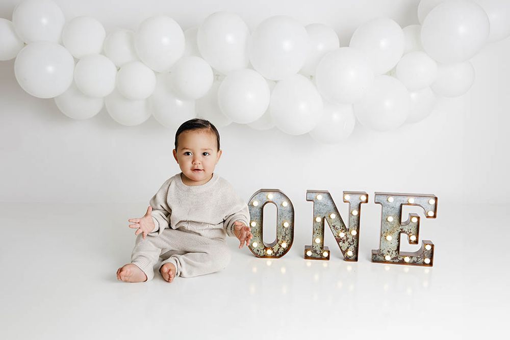 Baby boy in neutral outfit sits beside lit ONE marquee letters with white balloon backdrop for first birthday.