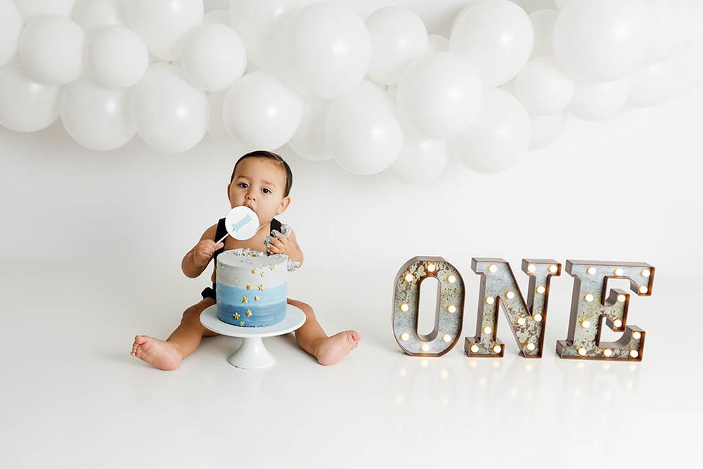Baby boy smashes first birthday cake next to lit ONE sign with white balloon backdrop by Winchester 1st birthday photographer.