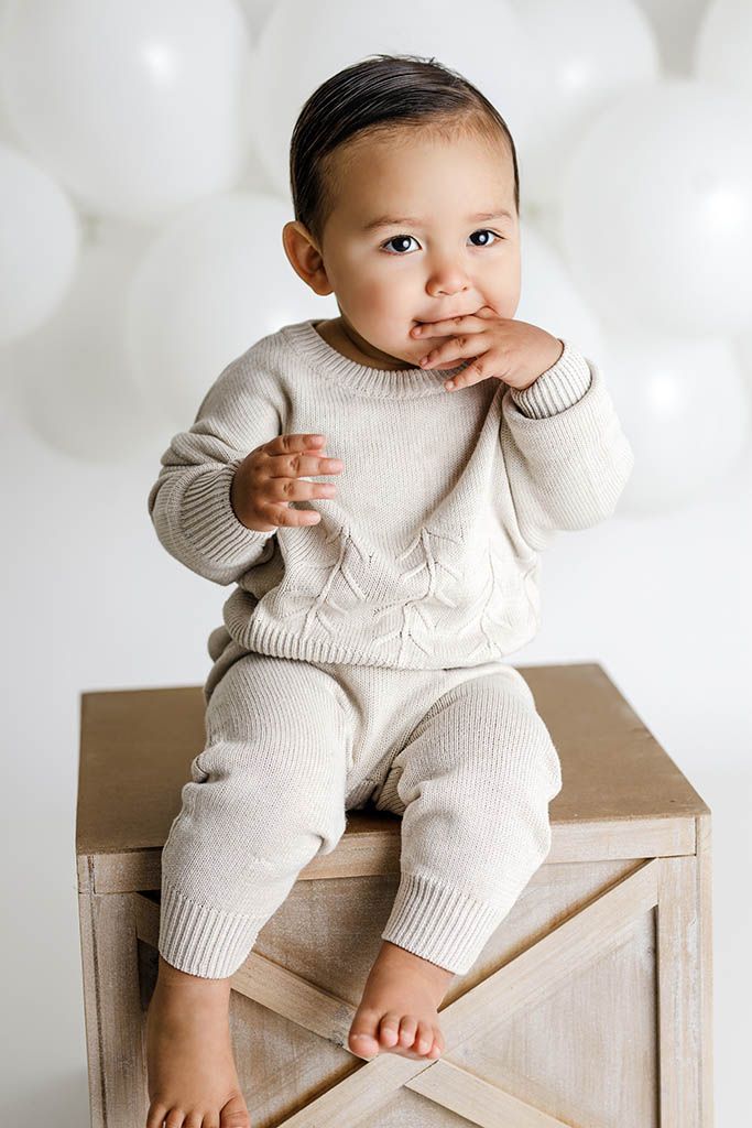 Adorable baby in cream knit sweater set sitting on wooden crate with white balloons in background.