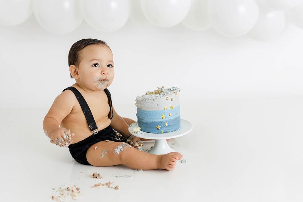 Winchester CA cake smash photographer - Baby boy in suspenders sits beside a blue smash cake during his first birthday photo session.