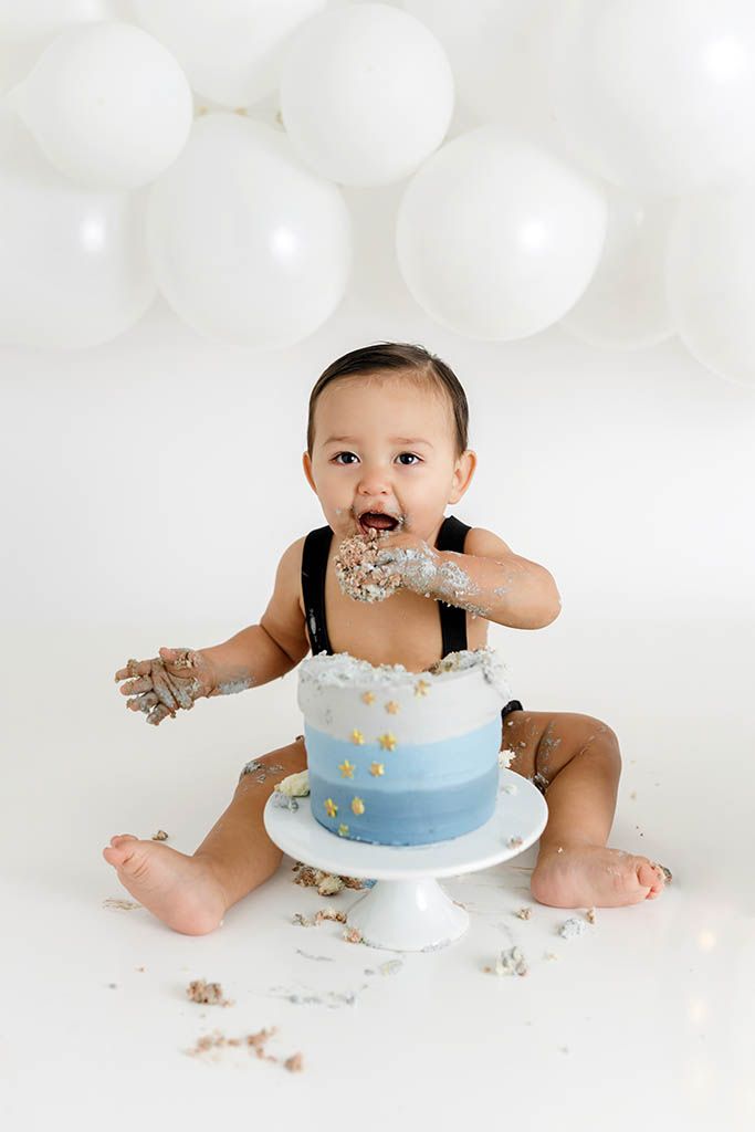 Adorable baby boy smashes first birthday cake surrounded by white balloons in studio photo session in Winchester CA.