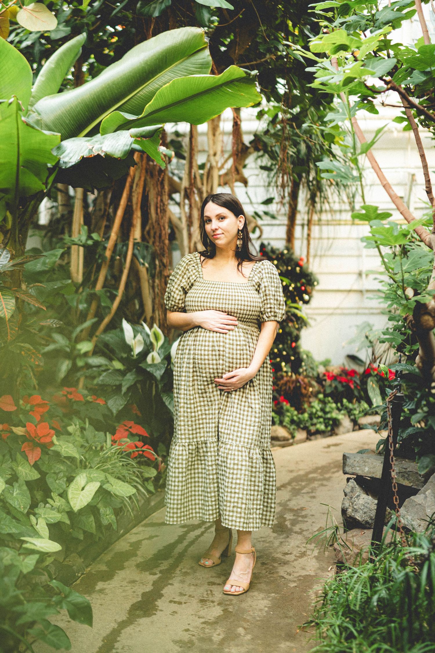 Pregnant woman in gingham dress cradling baby bump surrounded by lush tropical plants in a greenhouse garden.