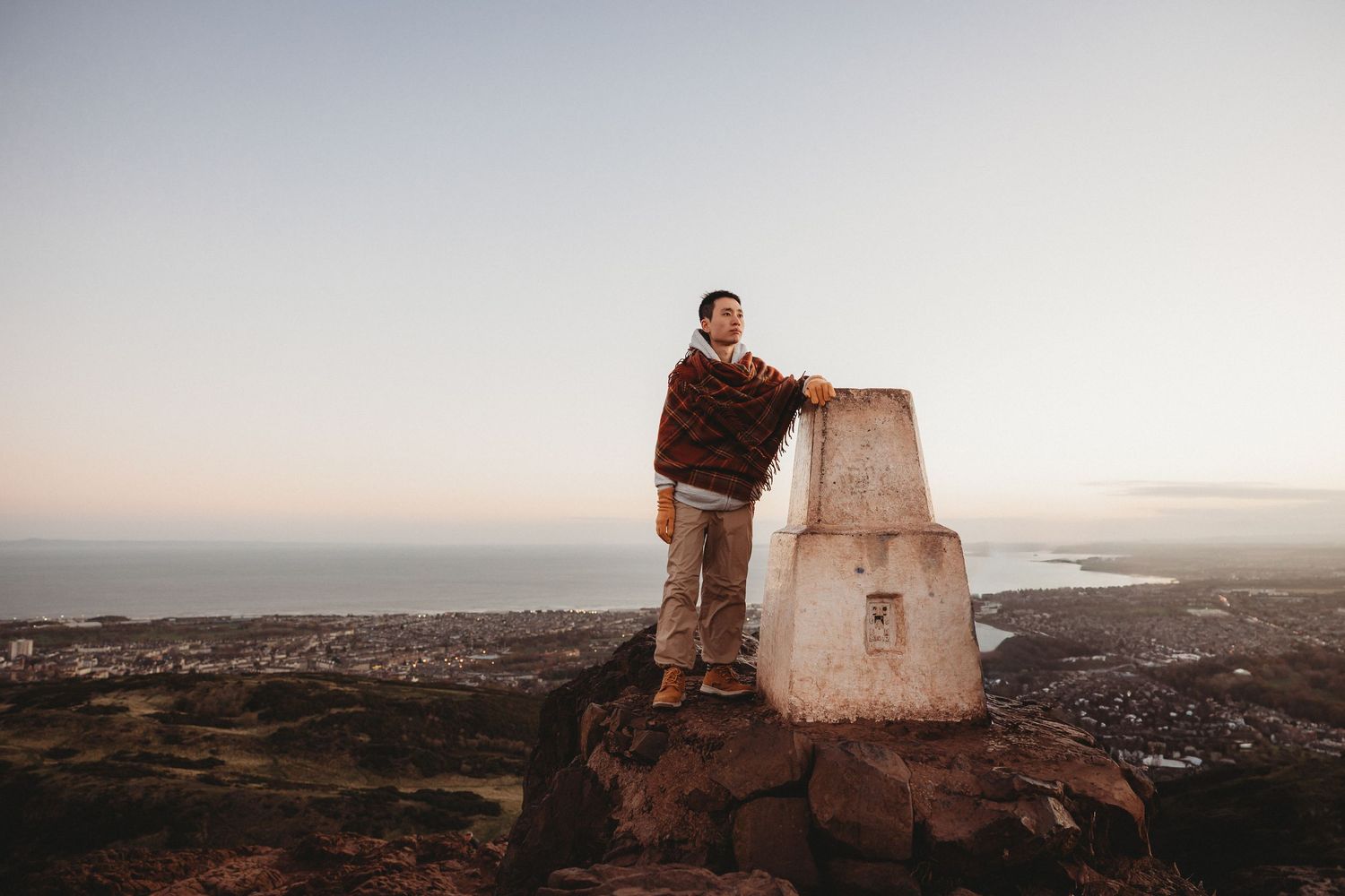 Person standing beside a trig point on a rocky hilltop at sunset with coastal city views below.