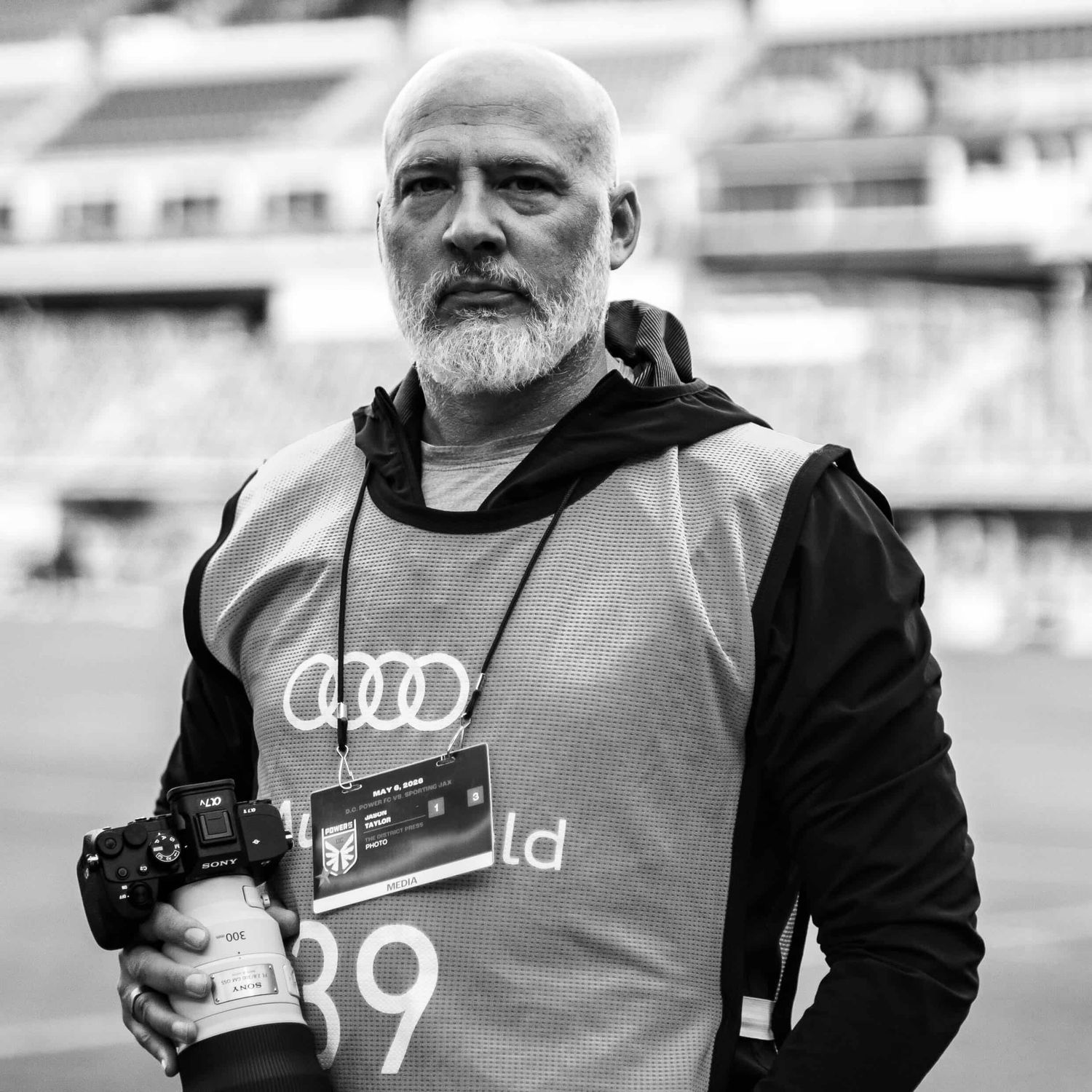 Bald man with white beard holding camera, wearing Audi press vest numbered 39 at a stadium, black and white photo.