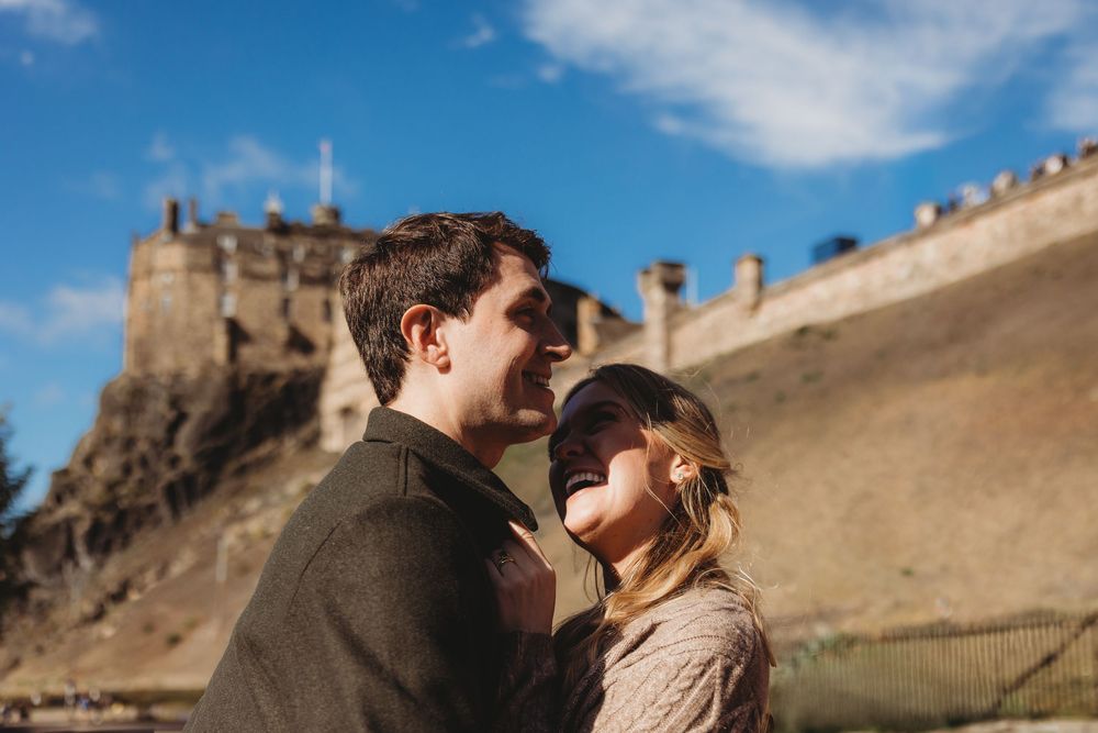 A couple embraces in front of Edinburgh Castle on a sunny day with blue skies and stone walls in the background.