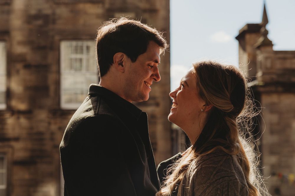 A couple in silhouette shares an intimate moment against a warm backlit sunset with brick walls in the background.