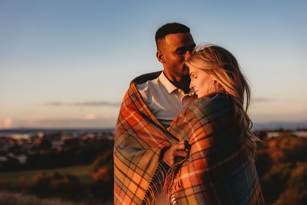 Couple wrapped in plaid blanket embraces during sunset at scenic coastal overlook.