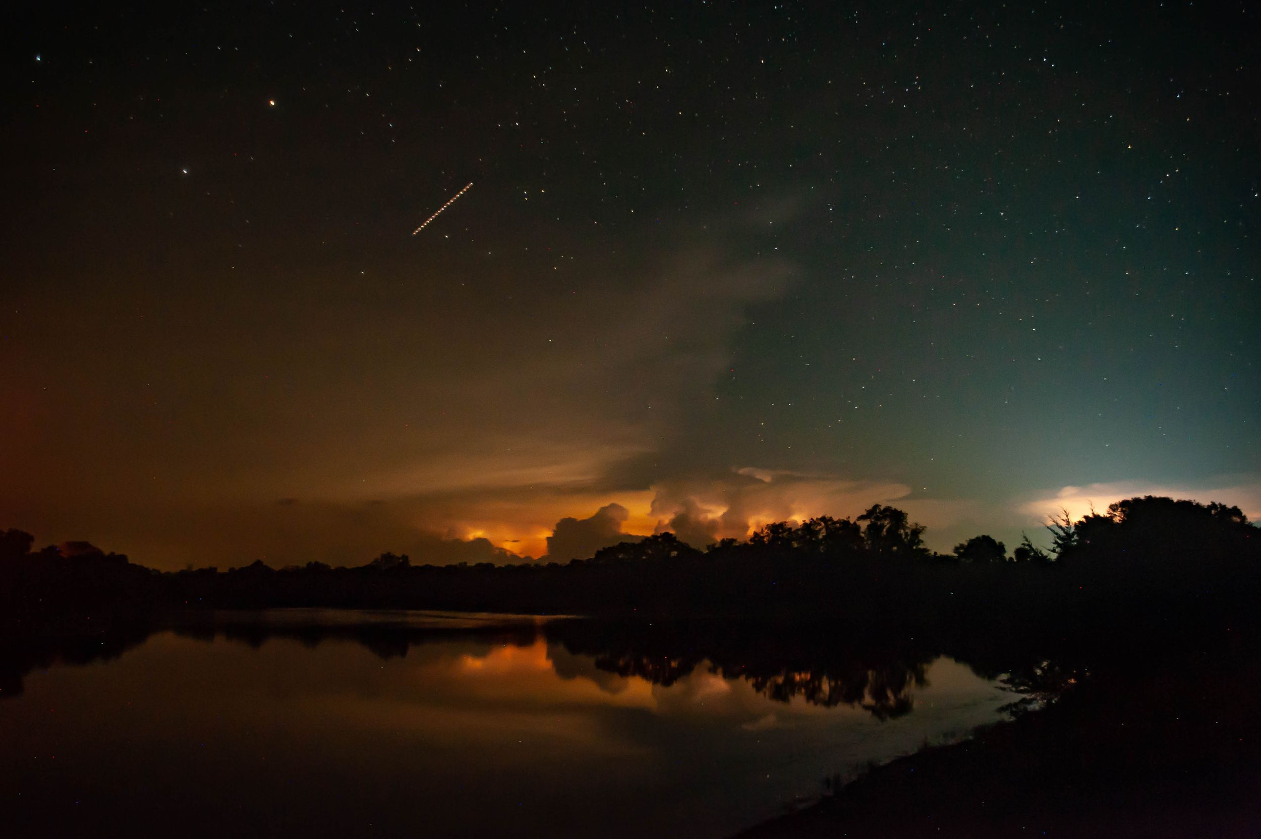 Night sky with satellite trail and lightning storm reflected in still lake Northern Missouri