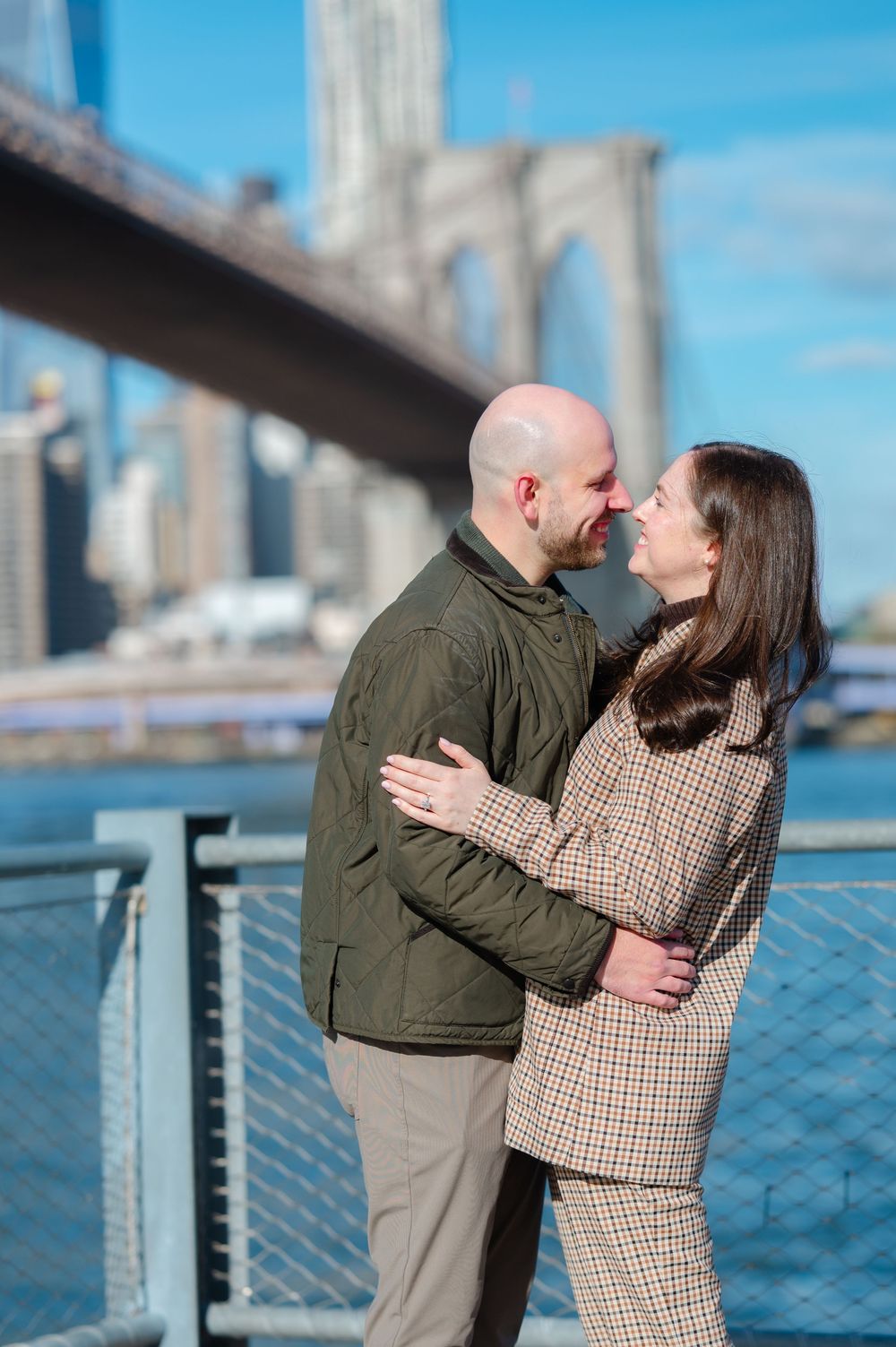 Couple embracing near Brooklyn Bridge on a sunny day, sharing a romantic moment by the waterfront.