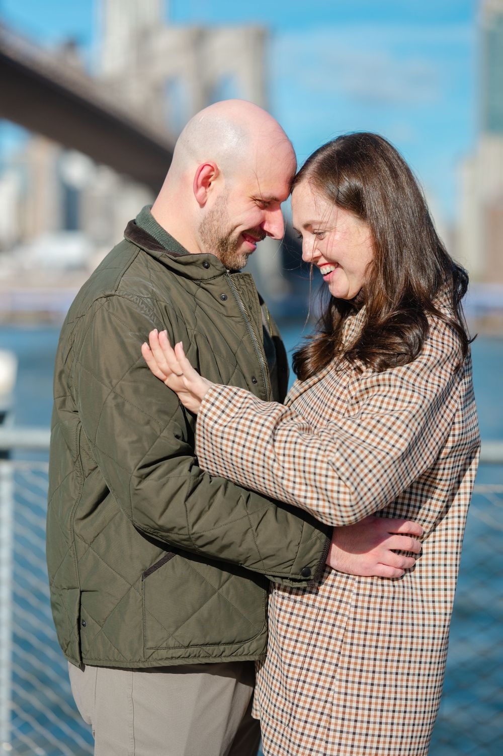 Couple embracing and smiling at each other near Brooklyn Bridge waterfront on a sunny day.