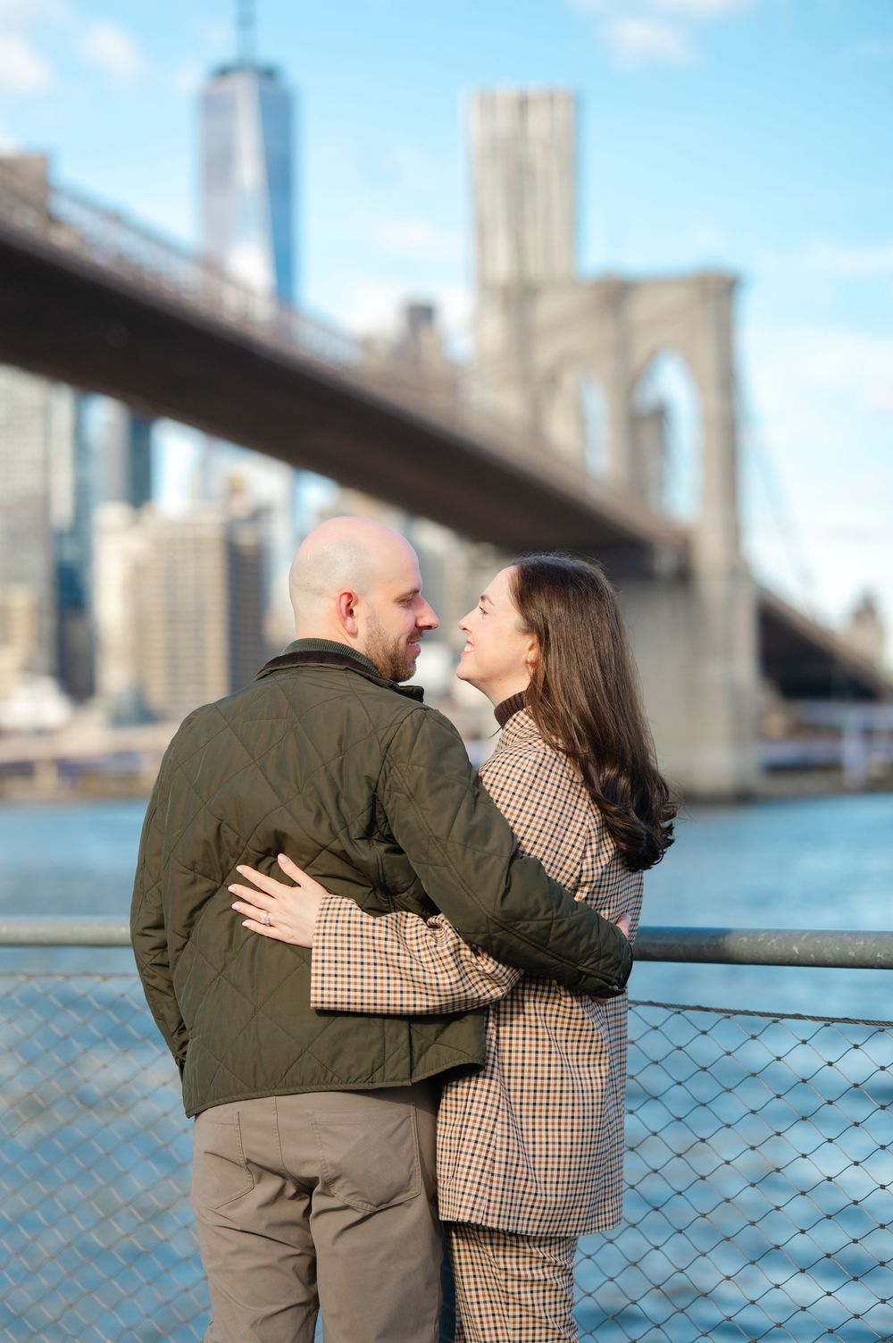 Couple embracing and smiling at each other on Brooklyn Bridge waterfront with Manhattan skyline behind.