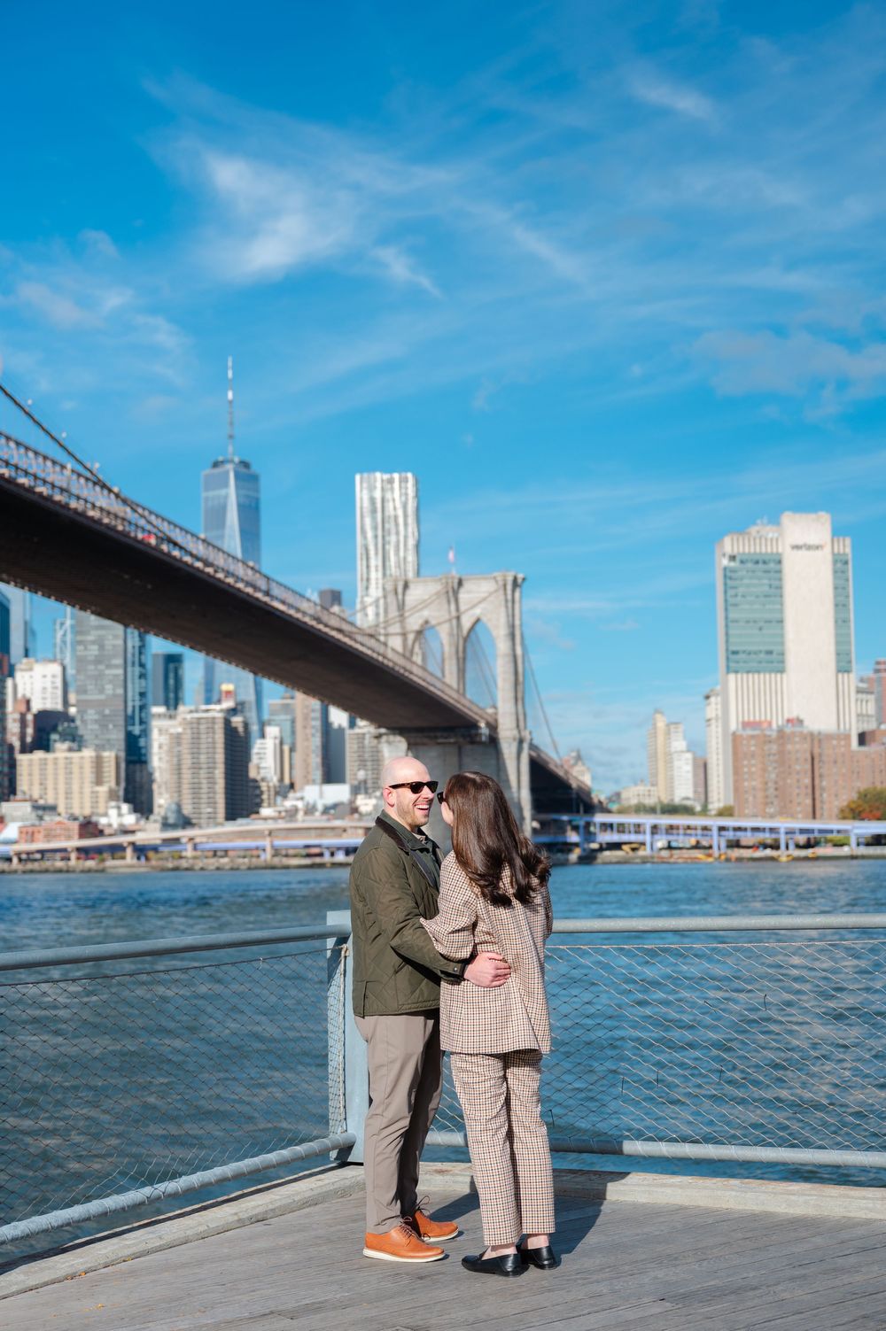 Couple embracing on a waterfront dock with the Brooklyn Bridge and NYC skyline behind them.