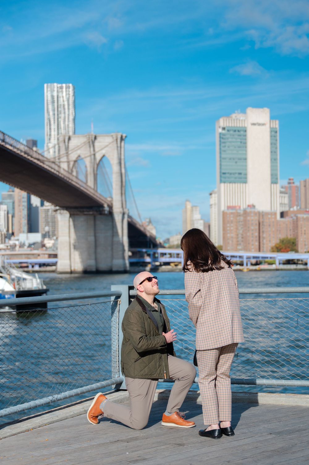 Man proposing on one knee to woman on Brooklyn Bridge waterfront pier with NYC skyline backdrop.