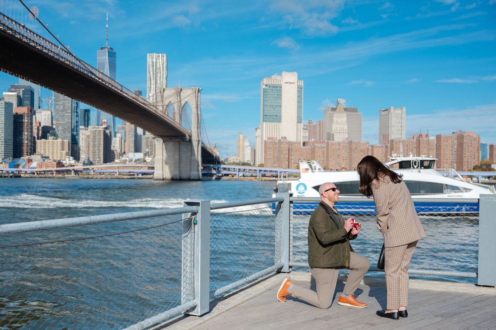 Man proposing on one knee to woman on Brooklyn Bridge Park waterfront with NYC skyline behind them.
