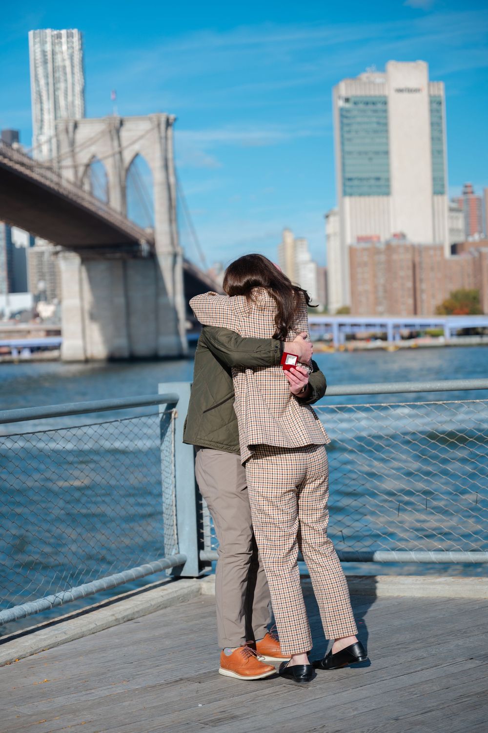 Couple embracing on a waterfront boardwalk near the Brooklyn Bridge after a marriage proposal in NYC.