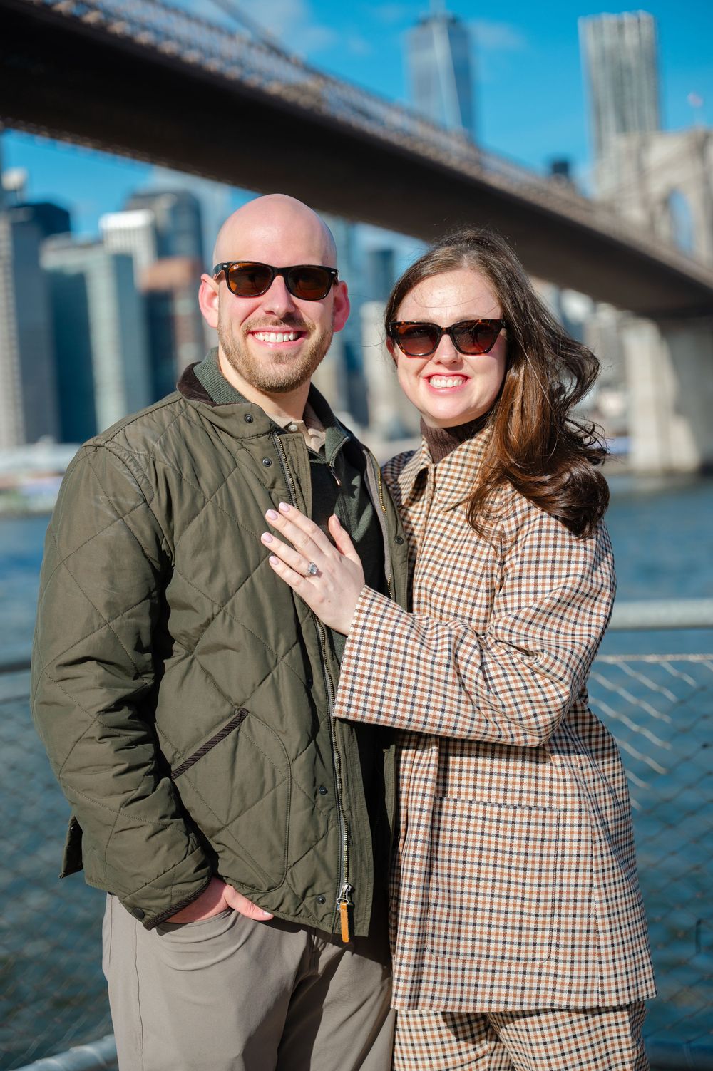 An engaged couple smiles outdoors near the Brooklyn Bridge, the woman displaying her engagement ring.
