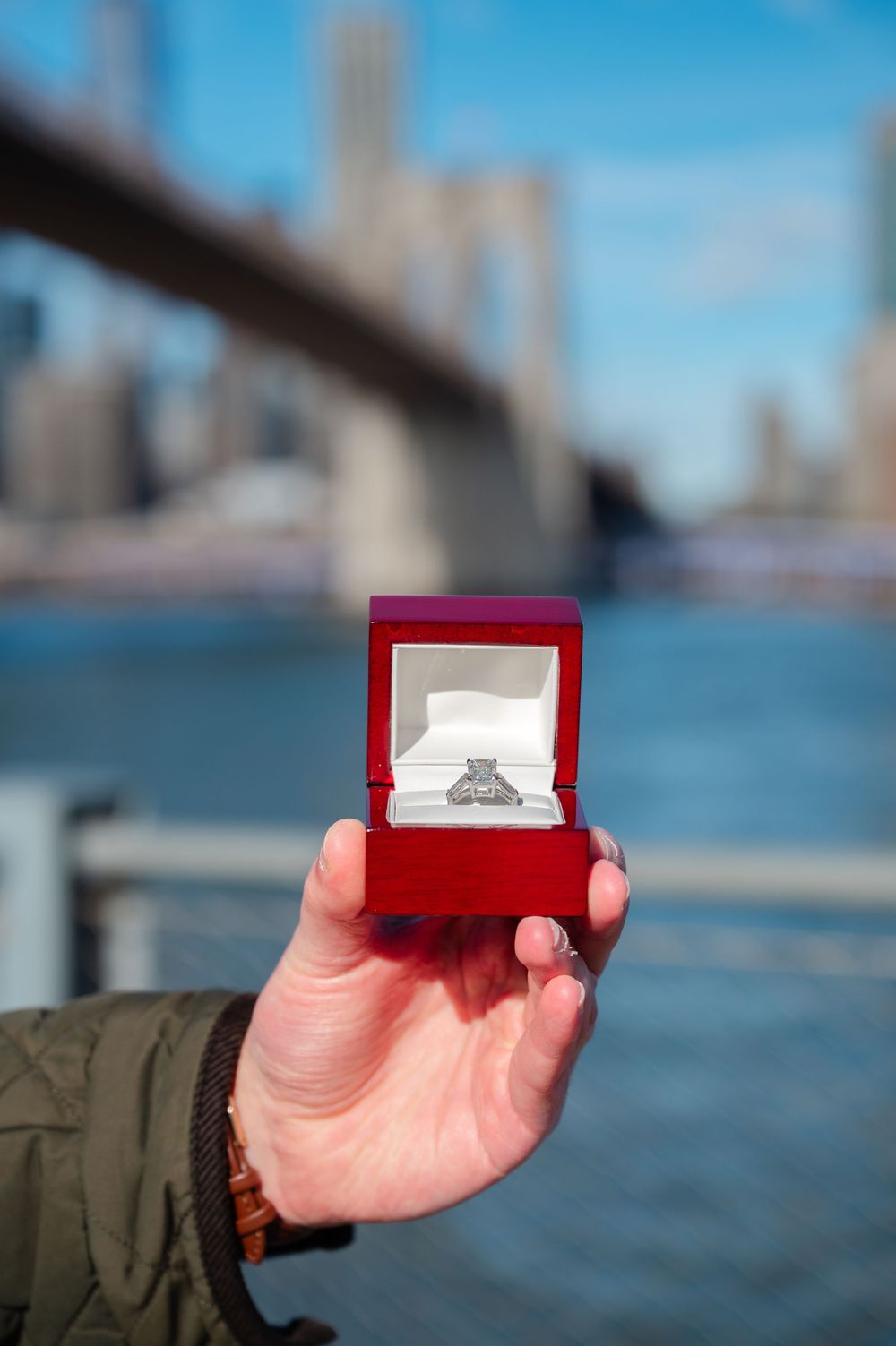 Hand holding red box with diamond engagement ring near Brooklyn Bridge in New York City on a sunny day.