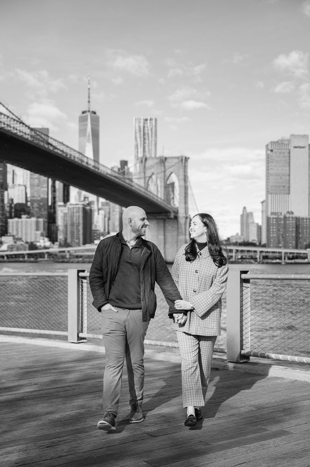 Black and white photo of a couple holding hands and walking along a dock with the Brooklyn Bridge behind them.