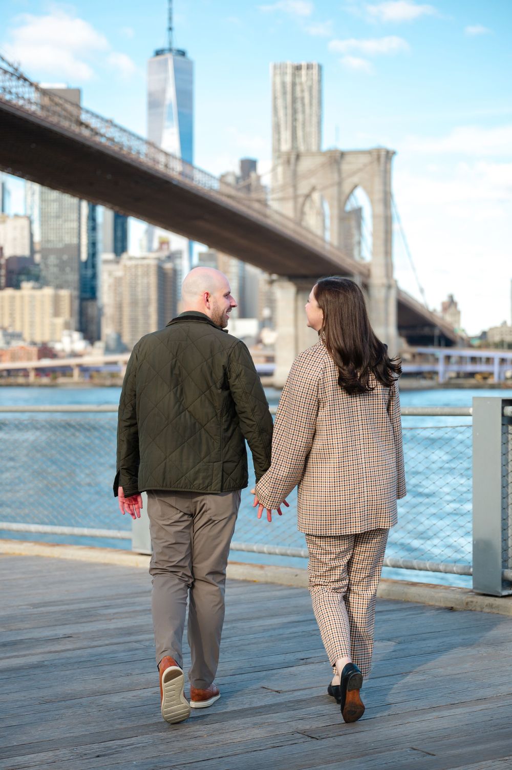 Engaged couple walks hand in hand along Brooklyn Bridge Park waterfront with NYC skyline backdrop.