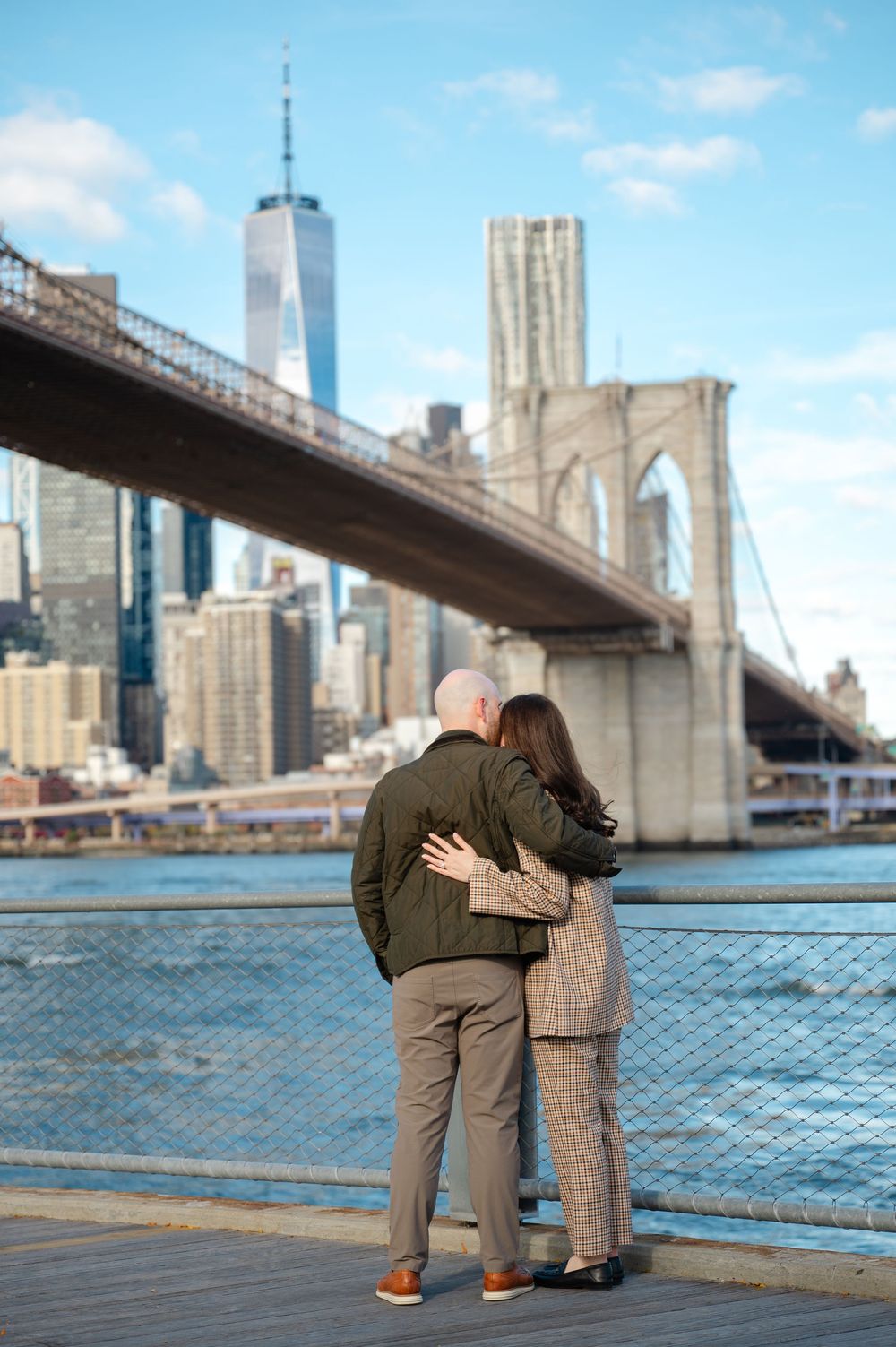 Couple standing arm in arm gazing at Brooklyn Bridge and Manhattan skyline from a wooden pier.