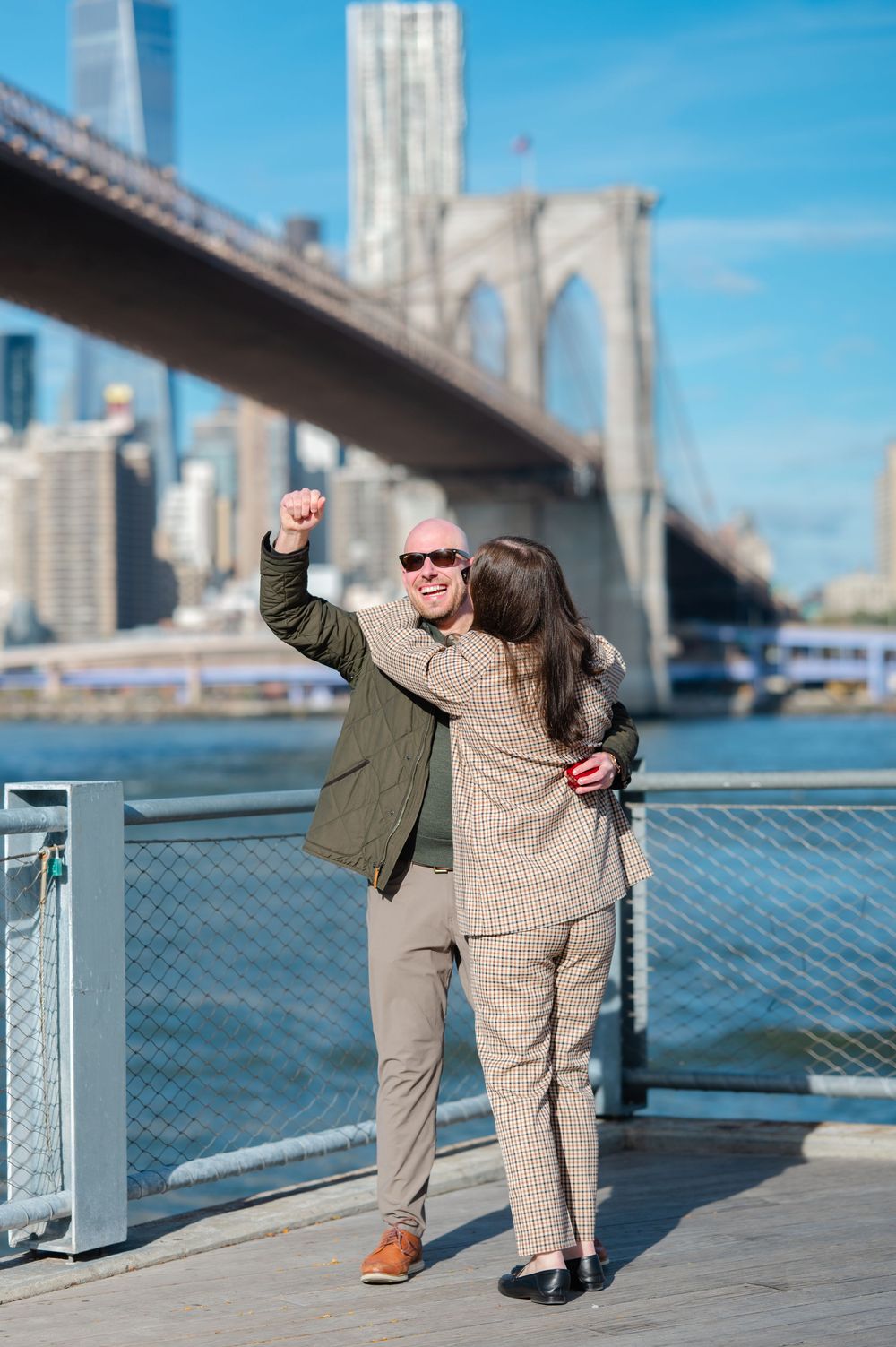 Engaged couple celebrating near Brooklyn Bridge waterfront, man raising fist while embracing woman in plaid suit.