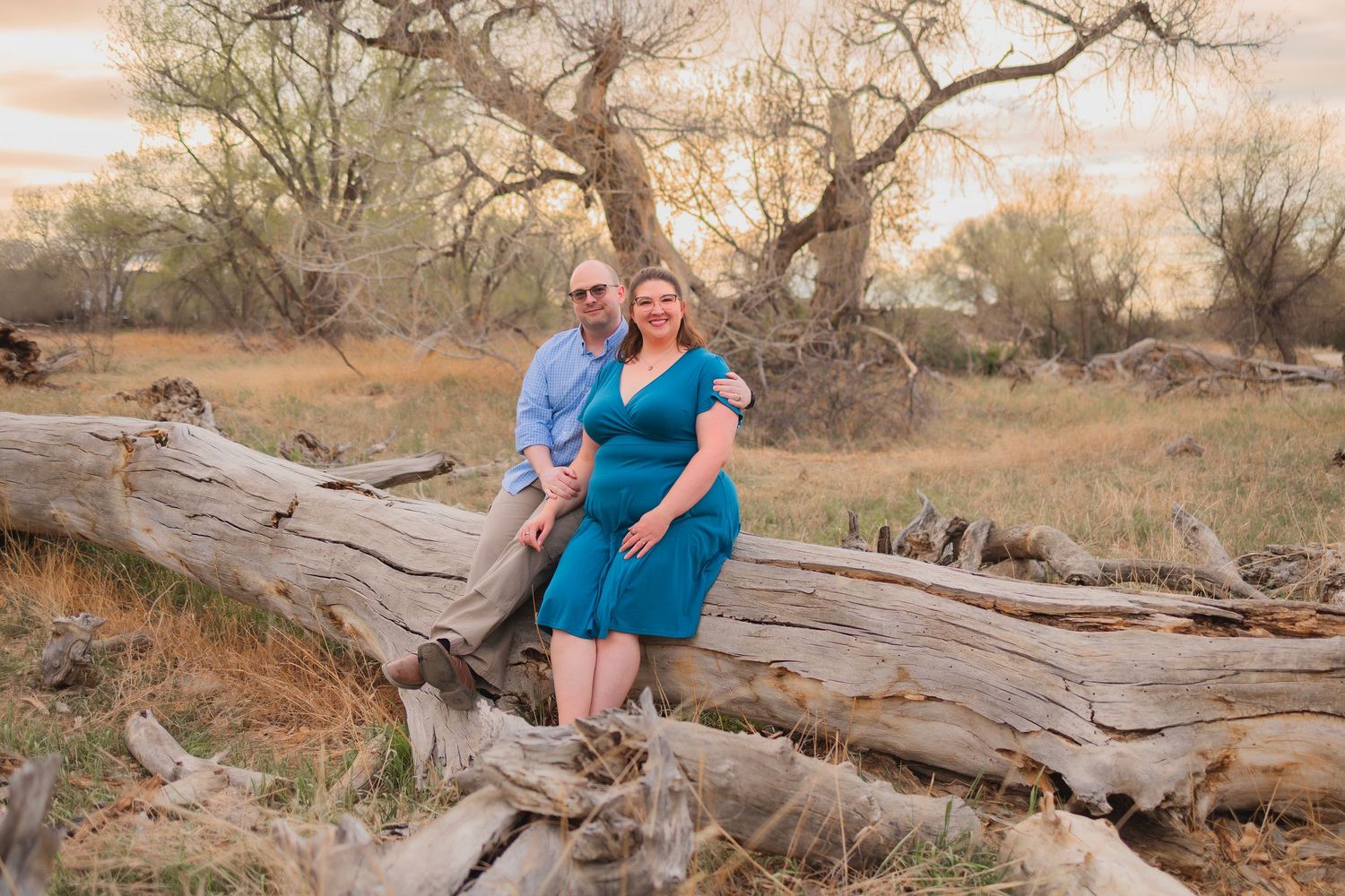 Couple posing on fallen log in rustic outdoor setting with golden grass and bare trees at sunset.