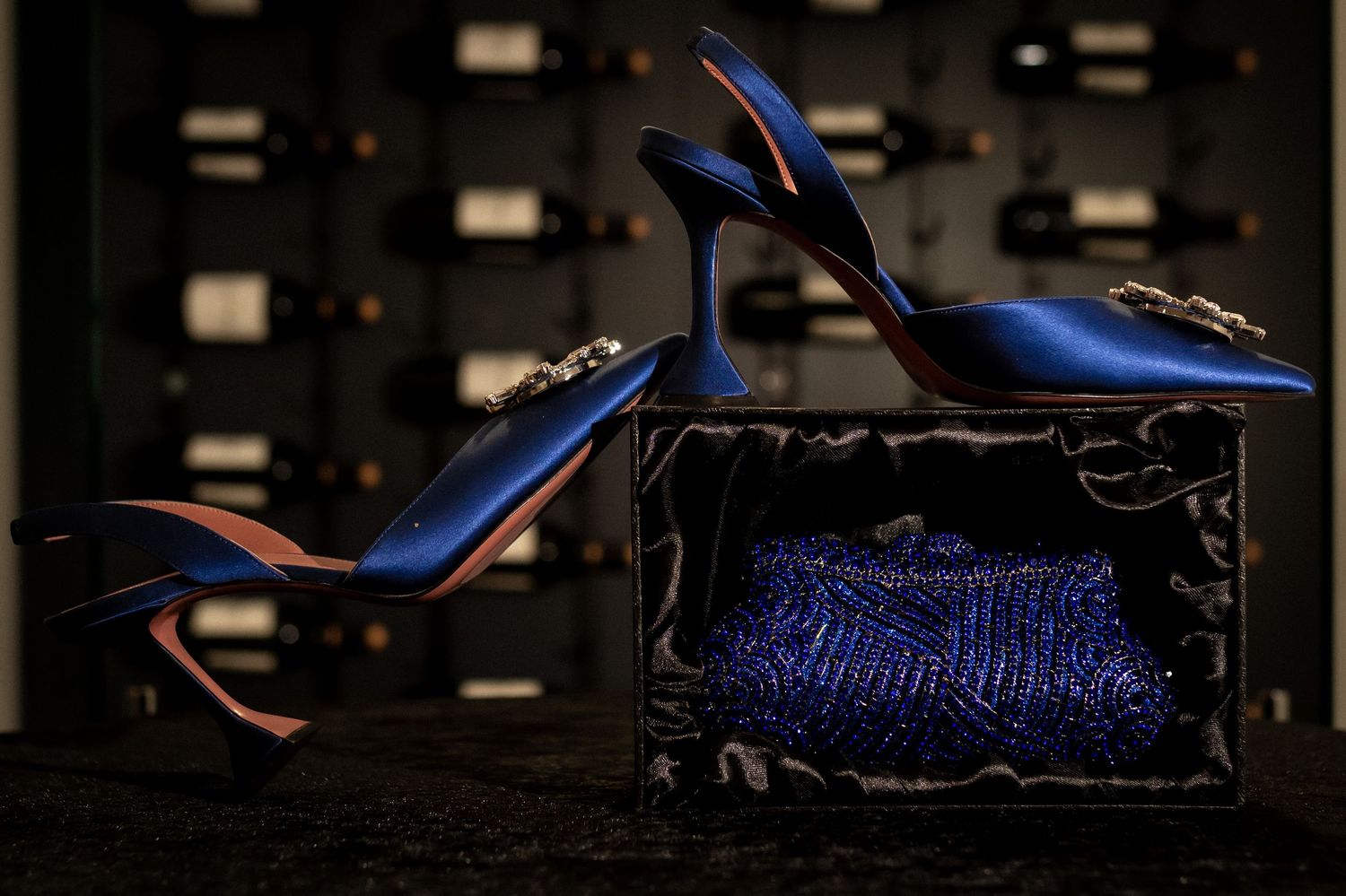 Elegant blue satin heels with crystal embellishments displayed on a matching blue sequined clutch in a wine cellar.